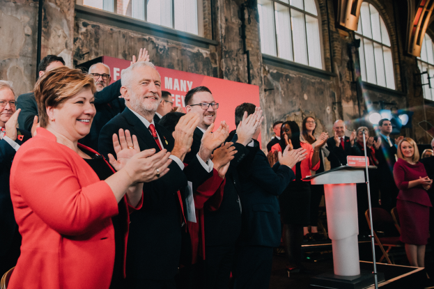 Eine Gruppe von Menschen, wahrscheinlich Liberale, steht vor einer Menge und klatscht feierlich, mit einem Podium, Mikrofon und Texttafel rechts, Stühlen, einer Fahne, einer Wand mit Fenstern und Lichtern im Hintergrund.