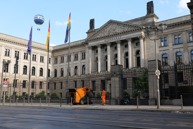 Großes Gebäude mit vielen Fenstern, als Bundestag in Berlin, Deutschland identifiziert, umgeben von Straßeninfrastruktur, Fußgängern und Flaggen, mit bewölktem Himmel im Hintergrund.
