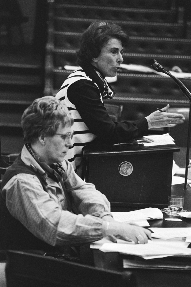 Schwarz-weißes Foto einer Frau, die an einem Tisch im Europäischen Parlament in Straßburg im Mai 1981 sitzt und einen Stift hält, mit Papieren, einem Glas Wasser und anderen Gegenständen auf dem Tisch, Treppe im Hintergrund.