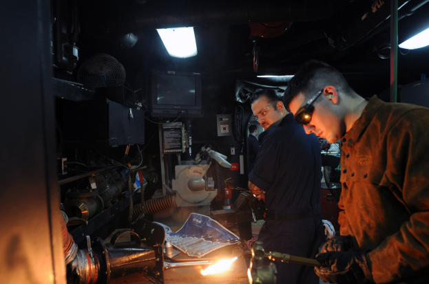 Zwei Männer mit Brille arbeiten an Metall in einer Werkstatt, einer verwendet ein Schweißgerät, während Werkzeuge und Equipment auf dem Tisch liegen, mit einem Fernseher und Lampen im Hintergrund.