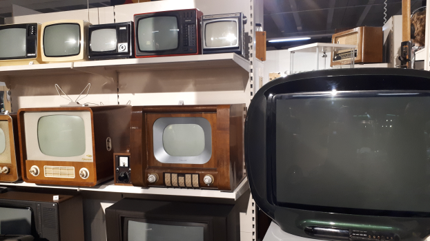 Ein Raum voller alter Fernseher auf Regalen, mit einer Wand im Hintergrund und einer Decke mit Lampen oben.