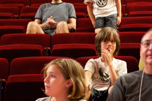 Ein Junge in einem weißen T-Shirt und schwarzen Hosen steht in der Mitte eines Kinosaals voller Menschen in roten Stühlen, umgeben von einer Atmosphäre der Vorfreude.