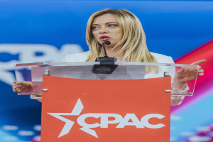 Eine Frau in einem weißen Kleid steht an einem Podium mit einem Mikrofon, die zu sein scheint eine politische Wissenschaftlerin, mit einem unscharfen Hintergrund.