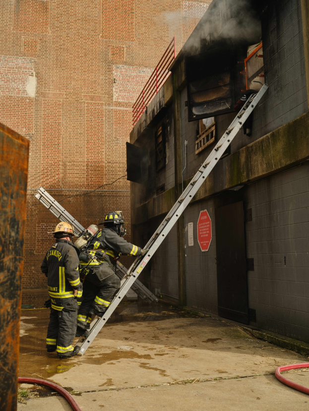 Feuerwehrleute mit Helmen klettern an einer Leiter zu einem raucherfüllten Gebäude hoch, mit einem Metallobjekt links und sichtbaren Rohren am Boden.