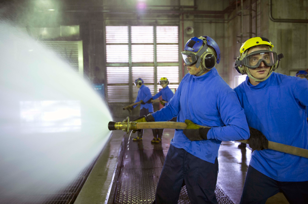 Eine Gruppe von Männern in blauen Hemden und gelben Helmen arbeitet an einer Maschine in einer Fabrikumgebung, wobei einer der Männer ein Rohr hält und Wasser auf den Boden sprüht.