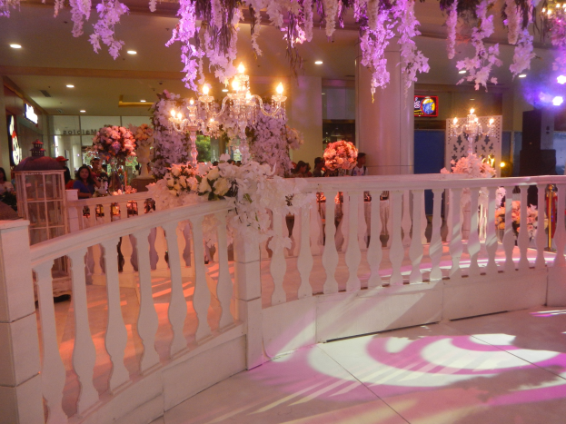 Eine Hochzeitsfeier mit einer weißen Balustrade, die mit Blumen geschmückt ist, Kronleuchtern an der Decke, Menschen, die sich darum versammeln, Säulen und Lichter im Hintergrund und ein Bildschirm auf der rechten Seite.