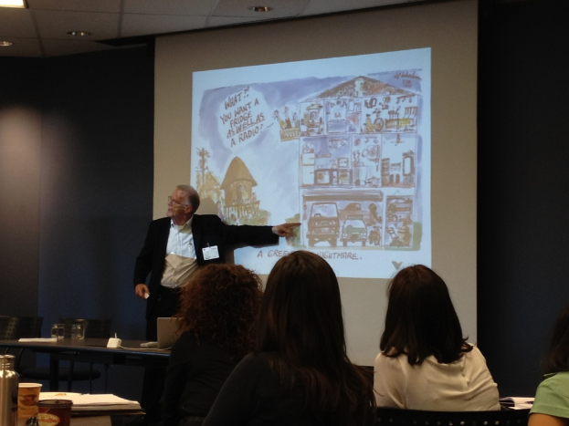 Ein Mann in einem weißen Hemd und schwarzer Jacke zeigt auf ein Fahrzeug auf einem Bildschirm in einem Konferenzsaal, mit drei Frauen vor ihm sitzend und einem Tisch mit einer Wasserflasche, einem Glas und einem Buch links.