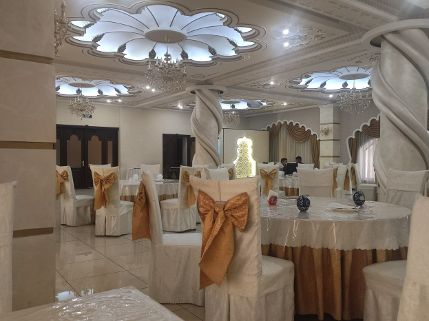 Ein großer Festsaal mit Tischen und Stühlen, die mit Tüchern bedeckt und mit verschiedenen Dekorationen geschmückt sind, mit Türen und Vorhängen an den Wänden und Kronleuchtern an der Decke.