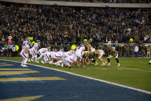 Ein Fußballspiel im Gange mit Spielern in Sportbekleidung und Helmen auf dem Feld, Zuschauern, Gittern und Tafeln mit Text im Hintergrund.