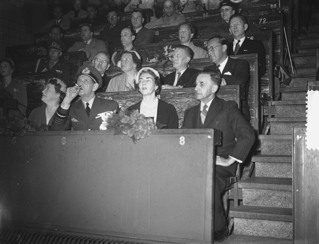Schwarzes und weißes Foto einer Zuschauermenge in einem Stadion, die einer Tribüne mit einem Blumenstrauß gegenüber sitzen, auf der rechten Seite sind Stufen sichtbar.