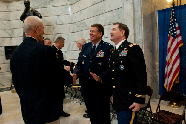 Menschen schütteln Hände mit Stühlen dahinter, eine Statue in der Nähe einer Wand im Hintergrund und eine Flagge und ein Vorhang auf der rechten Seite.