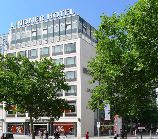 Großes modernes Hotelgebäude mit Glasfenstern und Beschilderung, umgeben von Bäumen und einer belebten Straße mit Fußgängern und Fahrzeugen, unter einem sichtbaren Himmel.