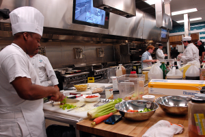 Eine Gruppe von Köchen in einer Küche, die Essen auf einem Schneidbrett zubereiten, mit einem Koch im Vordergrund in einer weißen Schürze und Mütze, der ein Messer hält, und umgeben von verschiedenen Utensilien, Schüsseln, Gemüse, einem Handy, Flaschen und anderen Gegenständen, mit einem Fernseher und Deckenlampen im Hintergrund.