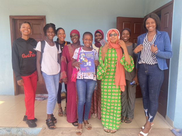 Eine Gruppe strahlender Frauen hält Bücher in den Händen und steht zusammen vor einem Schulgebäude in Nigeria, mit einem Tuch auf dem Boden und sichtbaren Türen im Hintergrund.