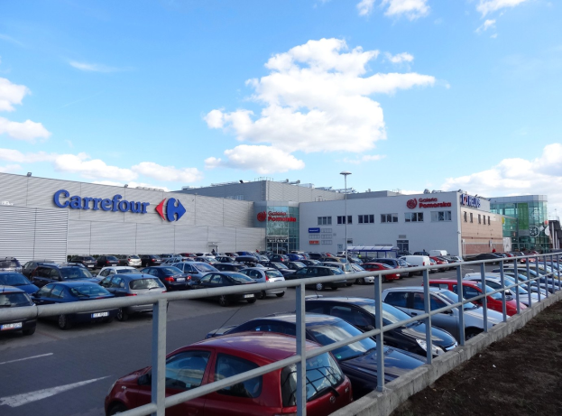 Großes Autohaus mit parkenden Fahrzeugen vorne, umgeben von Gebäuden, Straßeninfrastruktur und Fahrzeugen auf der Straße unter einem bewölkten Himmel.