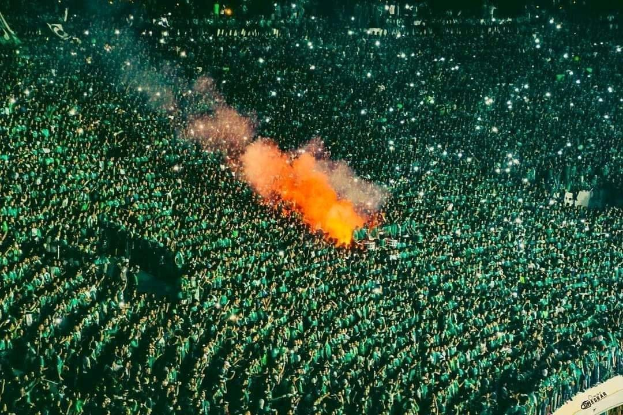Luftaufnahme einer großen Menge, die in einem Stadion steht und von einem zentralen Feuer beleuchtet wird.