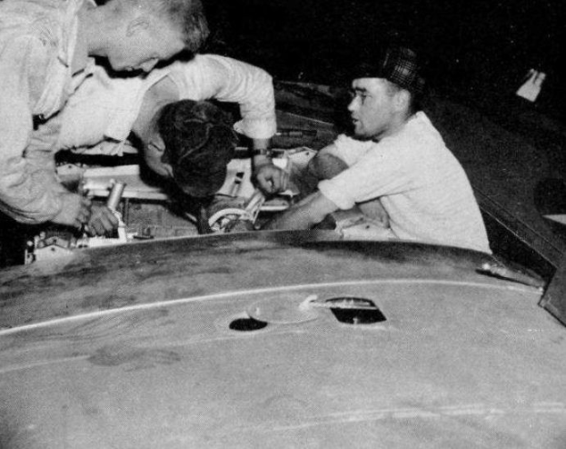 Drei Männer mit Mützen arbeiten an einem schwarz-weißen Auto in einer Garage, umgeben von Werkzeugen und Reparaturmaterialien.