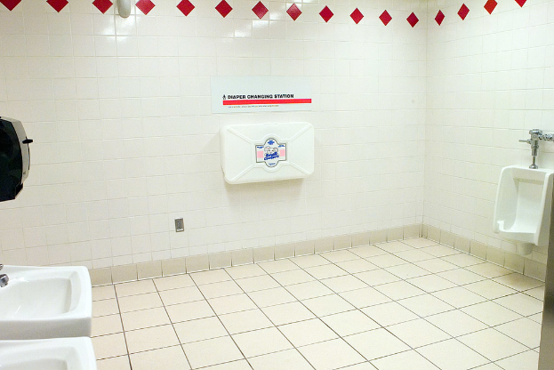 Ein Waschraum mit zwei Waschbecken im Vordergrund, eine Toilette auf der rechten Seite und eine Box vor gefliesten Wänden im Hintergrund.