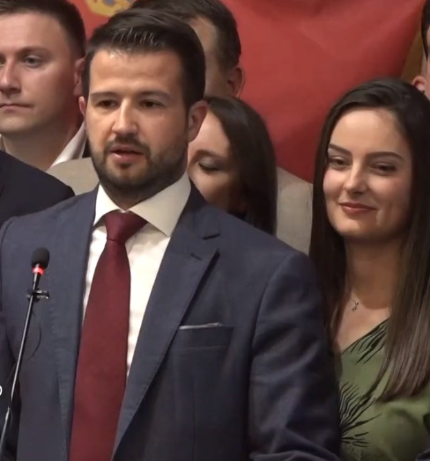 Ein Mann im Anzug spricht in ein Mikrofon auf einem Podium, mit einem Banner dahinter und einer Menge im Hintergrund, wahrscheinlich zu einem Verbot der sozialen Medien durch die Regierung.
