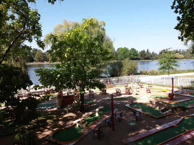 Minigolfanlage in einem Park mit grünem Rasen, Bäumen und einem Zaun, umgeben von verstreuten Stühlen und Tischen, mit einem Wasserlauf und einem Boot im Hintergrund unter einem sichtbaren Himmel.