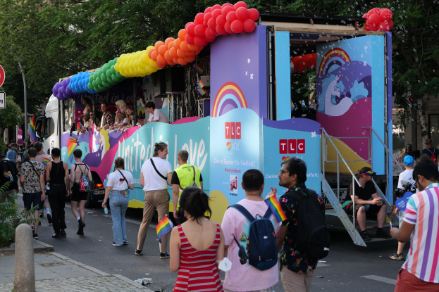 Eine Gruppe von Menschen geht neben einem Lastwagen mit bunten Luftballons die Straße entlang, mit Schildern an der Straße und Bäumen und Gebäuden im Hintergrund, was auf eine Pride-Parade in Paris hindeutet.