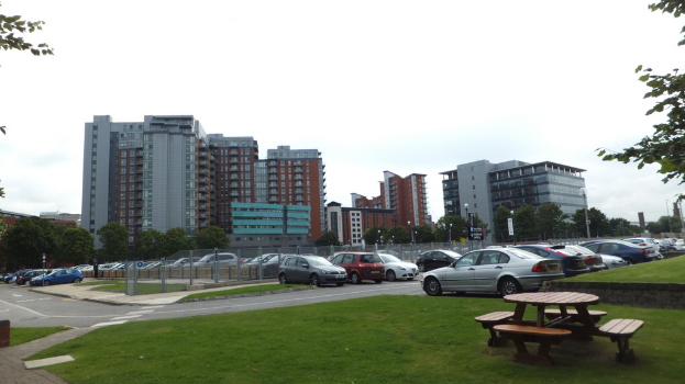 Ein Parkplatz mit zahlreichen Autos, umgeben von Gras, Bäumen und Gebäuden, unter einem klaren Himmel, mit einer Bank und Stühlen auf dem Rasen.