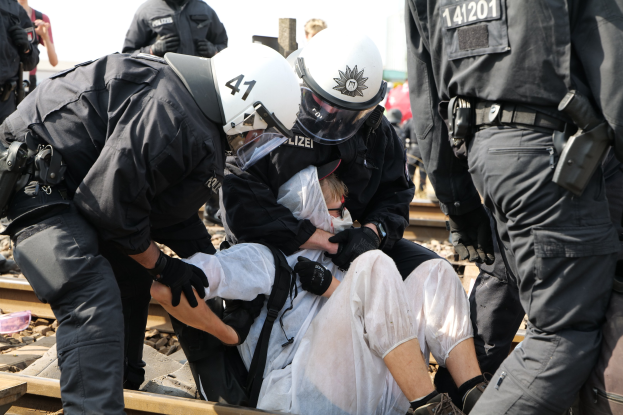 Eine Gruppe von Polizisten in Einsatzausrüstung verhaftet einen Mann in einem weißen Kleid auf einem Bahngleis, mit anderen im Hintergrund sichtbar.