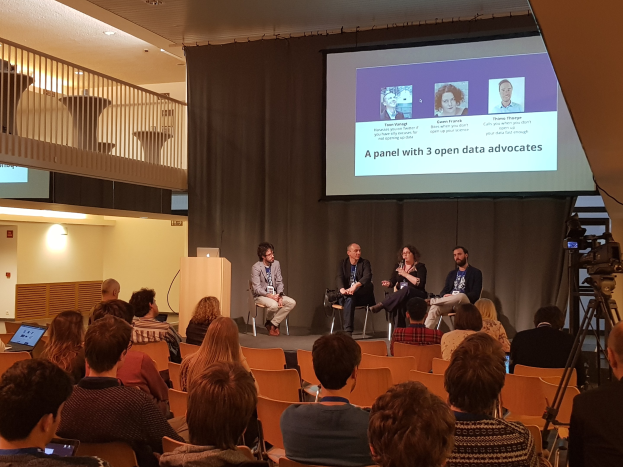 Eine Gruppe von Menschen sitzt auf Stühlen vor einem Projektionsschirm mit einem Laptop auf einem Tisch, einer Kamera auf einem Stativ rechts daneben und Geländern, Lampen und einer Wand im Hintergrund, die an einer Diskussion über Open-Data-Vertreter teilnehmen.