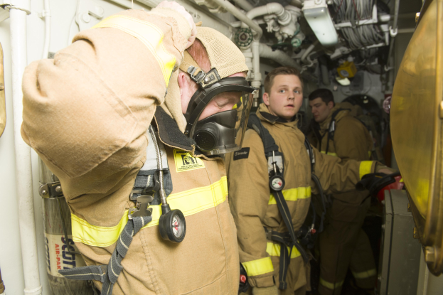 Feuerwehrleute in Gasmaske und Schutzausru├čtung stehen in einem Raum mit Rohren und Gegenst├Ąnden, mit einem Metallgegenstand rechts.