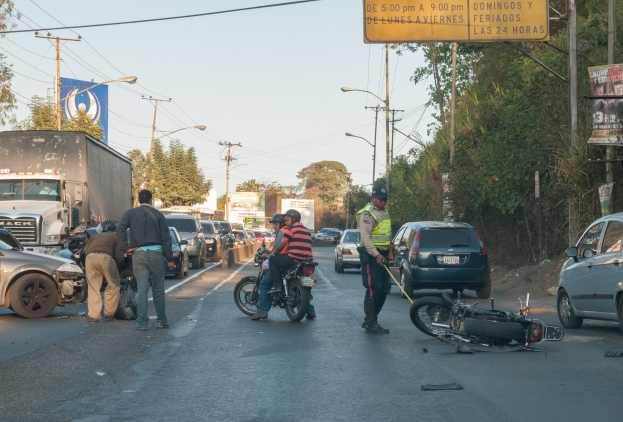 Gruppe von Menschen um ein verunglücktes Motorrad am Straßenrand mit mehreren Fahrzeugen, einschließlich eines Lastwagens, im Hintergrund und Bäumen, Pfosten, Laternen, Schildern und Himmel im Bild.