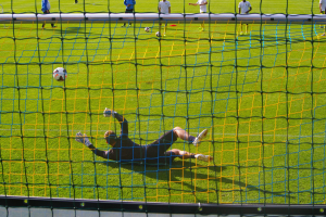 Ein Fußballspieler hält vor einem Tor, mit einem Netz im Vordergrund und einigen Menschen im Hintergrund, während mehrere Fußballschuhe auf dem Boden verstreut sind.