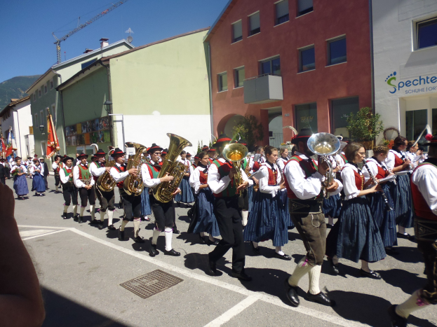 Eine Gruppe von Menschen in traditioneller bayrischer Tracht, die auf der Straße Musikinstrumente spielen, während sie an Gebäuden vorbeigehen, einige halten Fahnen, mit einem Hügel und einem blauen Himmel im Hintergrund.