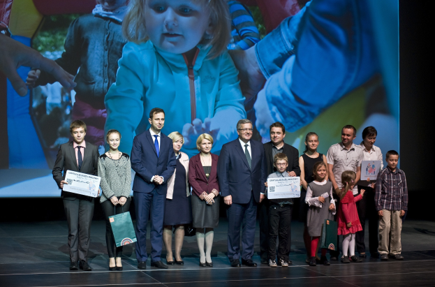 Eine Gruppe von Menschen auf einer Bühne, einige halten Taschen und Urkunden, mit einem Bildschirm im Hintergrund, der ein Bild eines Kindes und anderer Menschen anzeigt.