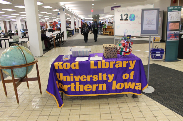 Eine Ausstellung mit einem Tisch, auf dem verschiedene Landesflaggen, eine Kiste, Papiere und ein Banner stehen, eine Kugel zur Linken, ein Anschlagbrett zur Rechten mit einigen Leuten in der Nähe und mehrere Tische und Stühle in der Ferne.