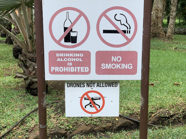 Schild am Straßenrand mit dem Text "Alkoholgenuss ist Verboten" und "Rauchen Verboten", mit Bäumen und Gras im Hintergrund.