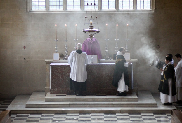 Eine Statue steht in der Mitte mit Kerzen auf Haltern, umgeben von Menschen am Boden, mit einer Wand und Fenstern im Hintergrund.