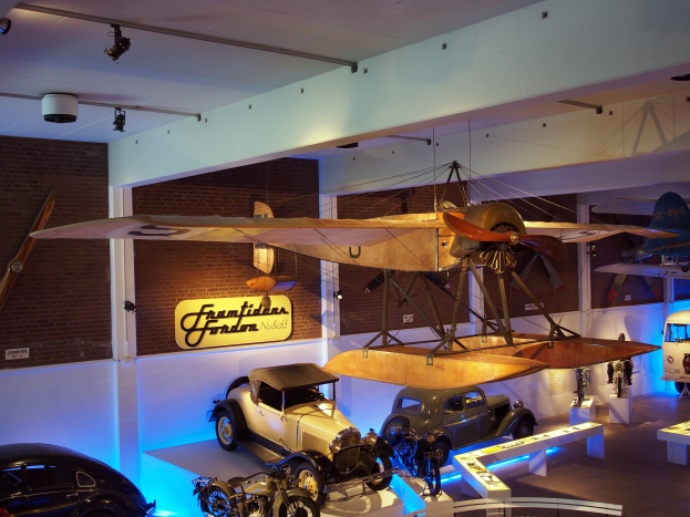 Eine Museumsausstellung, die verschiedene Autos, Motorräder und ein Flugzeug zeigt, das von der Decke hängt, mit Informationsschildern an den Wänden und Deckenbeleuchtung.