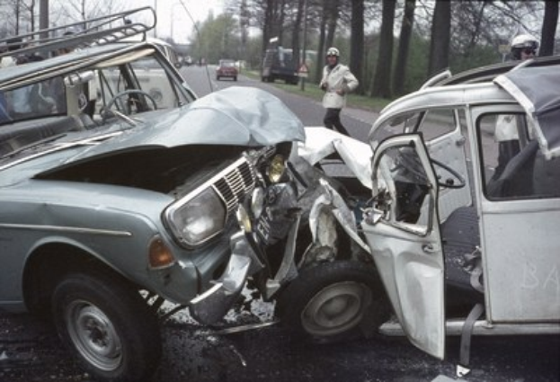Ein verunglücktes Auto am Straßenrand mit Zuschauern in Helmen und anderen Fahrzeugen im Hintergrund, vor einem Baum, Pföhlen und einem klaren blauen Himmel.