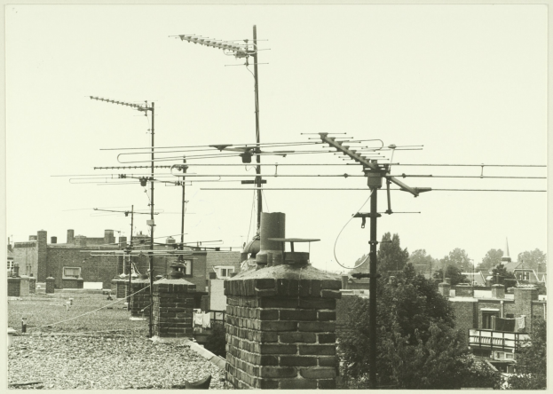 Ein Schwarz-Weiß-Foto einer Stadtkulisse mit zahlreichen Gebäuden, Bäumen und Fernsehantennen auf einem Dach.