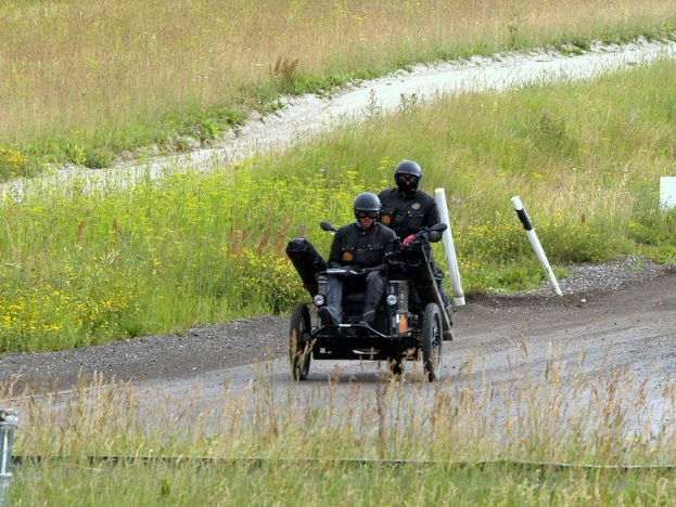 Zwei Polizisten in schwarzen Uniformen und Helmen fahren auf einem dreirädrigen Motorrad über einen von Gras und Pflanzen gesäumten Schotterweg, mit weißen Pfählen im Hintergrund.