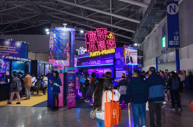 Eine große Gruppe von Menschen vor einem Stand auf einer Messe, einige halten Taschen und Schilder mit der Aufschrift 'Indie Game Development Award 2019' im Hintergrund.
