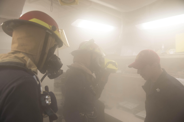 Feuerwehrleute in Helmen und Ausrüstung atmen Rauch während eines Trainings, mit einem, der eine Kamera hält und Hintergrundlichter sichtbar sind.
