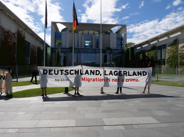 Eine Gruppe von Menschen mit einem Banner 'Deutschland, Lagerland Migration ist kein Verbrechen' vor einem umzäunten Gebäude mit Bäumen und Flaggen.