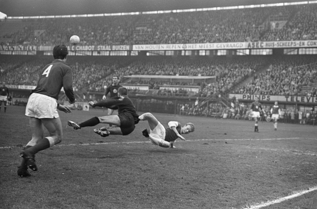 Eine Gruppe von Männern, die auf einem Feld in einem Stadion Fussball spielen, mit Zuschauern auf den Tribünen und Bannern im Hintergrund, dargestellt in Schwarz-Weiß.