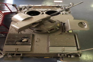 Ein großer Militärpanzer mit sichtbaren Ketten und Turm sitzt auf einem Boden, mit einem Metallgeländer auf der linken Seite.