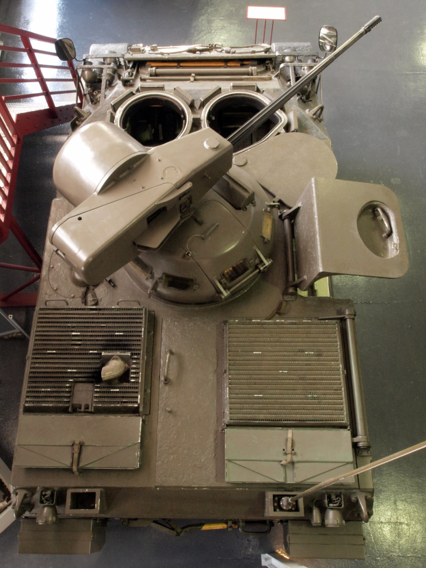 Ein großer Militärpanzer mit sichtbaren Ketten und Turm sitzt auf einem Boden, mit einem Metallgeländer auf der linken Seite.