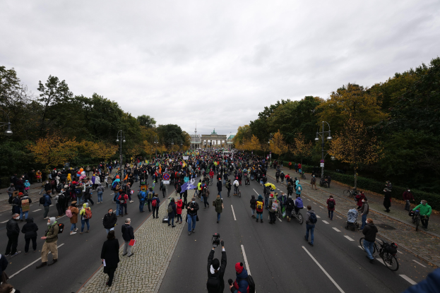 Große Gruppe von Menschen, die eine von Bäumen gesäumte Straße mit Laternenmasten entlanggehen und Kameras halten, während sie an einer Protestkundgebung in Berlin teilnehmen, mit einem Gebäude und einem klaren Himmel im Hintergrund.