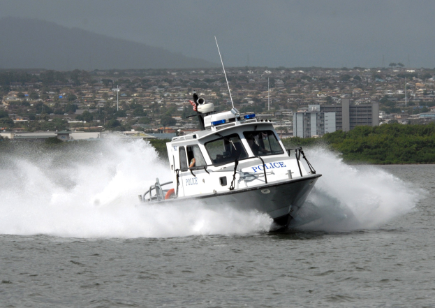 Polizeiboot fährt durch Wasser in der Nähe einer Stadt mit Passagieren an Bord, umgeben von Bäumen, Gebäuden, Strommasten, Bergen und einem klaren Himmel.