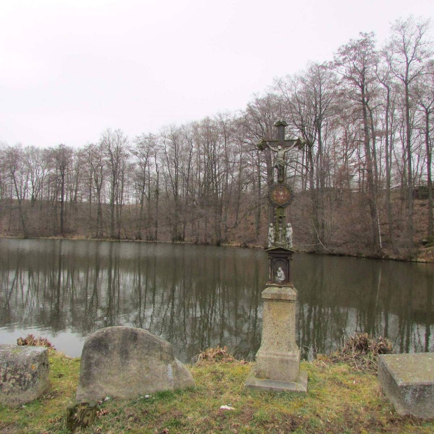 Ein Friedhof mit einem Kreuz in der Mitte eines Sees umgeben von Bäumen, saftigem Grün, verstreuten Grabsteinen und einem sichtbaren Himmel im Hintergrund.