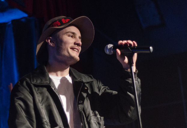 Ein Mann in einer schwarzen Jacke und Hut lächelt, während er ein Mikrofon vor sich hält.